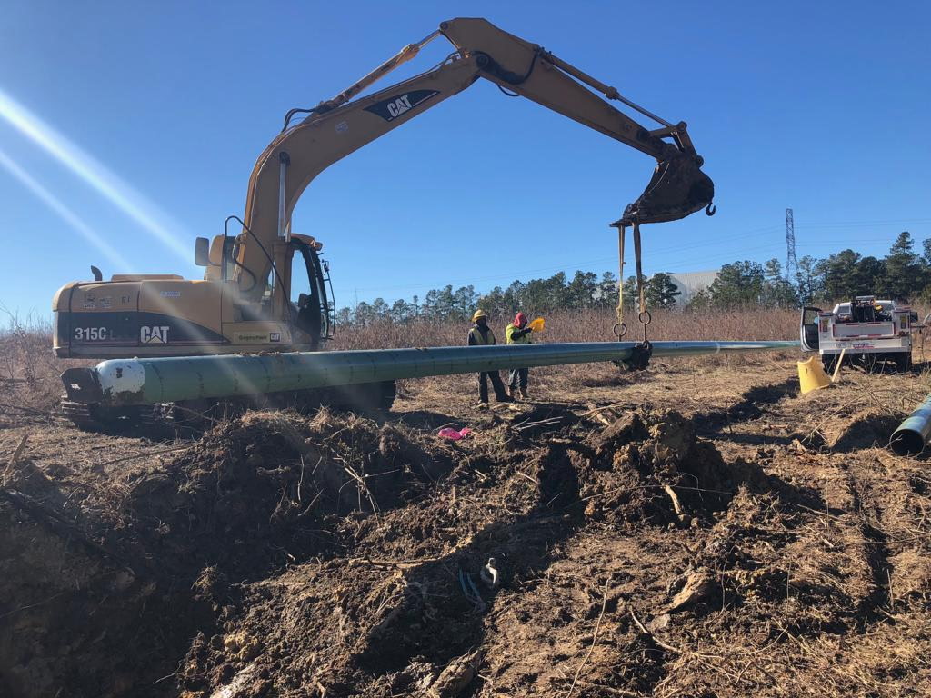 Excavator setting a large-diameter steel casing pipe while a Triple J Specialist crew guides it into place